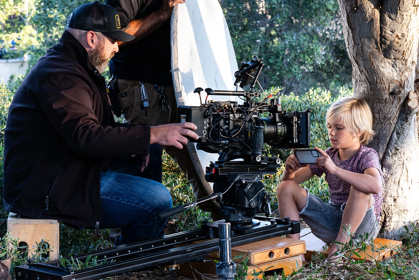 A film set shows a camera on a dolly filming a close up of a young blonde boy on a phone