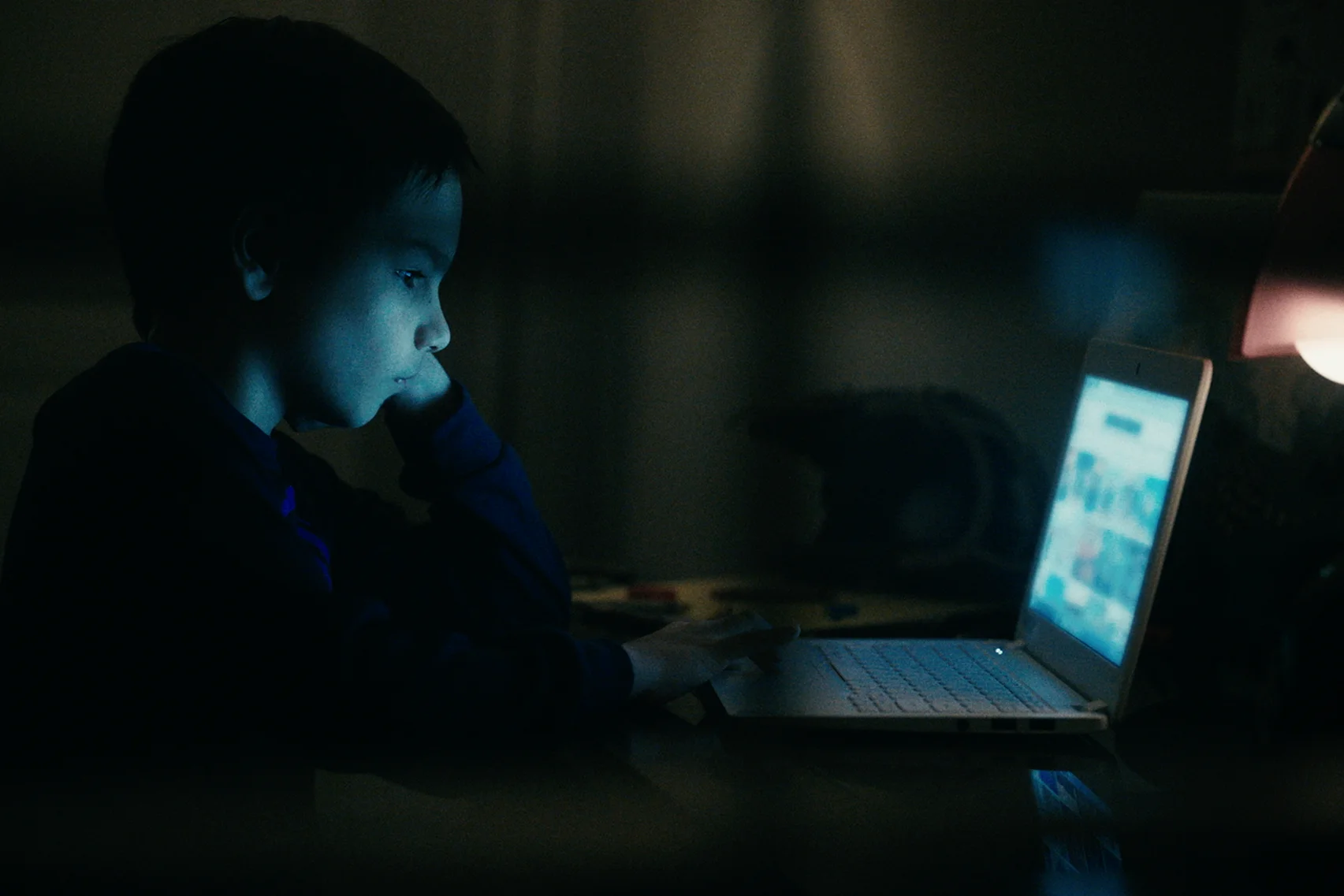 A young boy is sitting at a desk, looking at a blurry laptop in a dark room