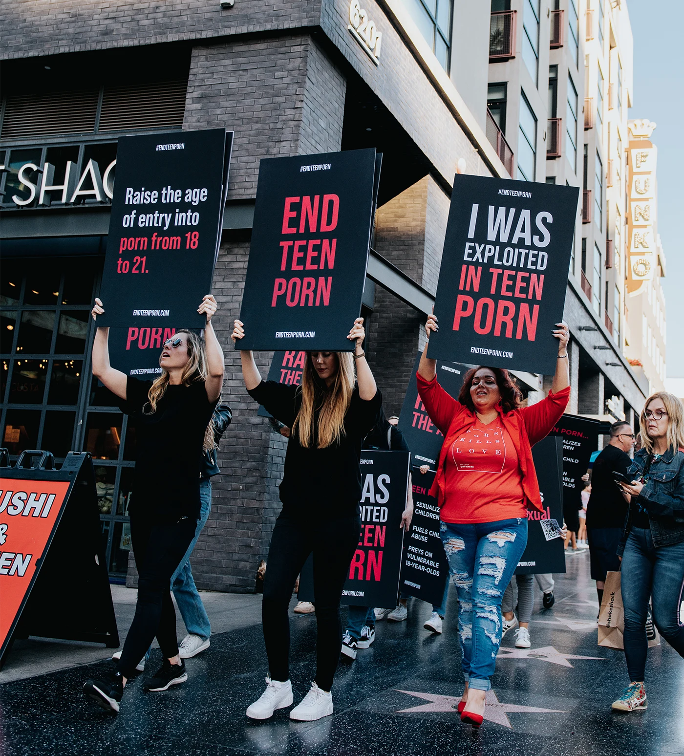 Three women hold up signs in protest that say things like End Teen Porn and I Was Exploited in Teen Porn